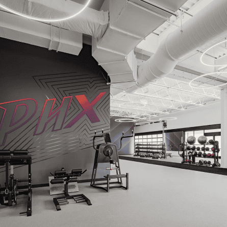Modern gym interior with weight racks, exercise machines, and a large PHX wall graphic.