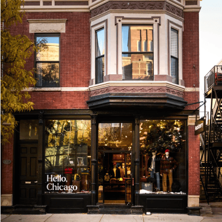 Red-brick Chicago storefront with bay windows, mannequins, and a window sign reading “Hello, Chicago”