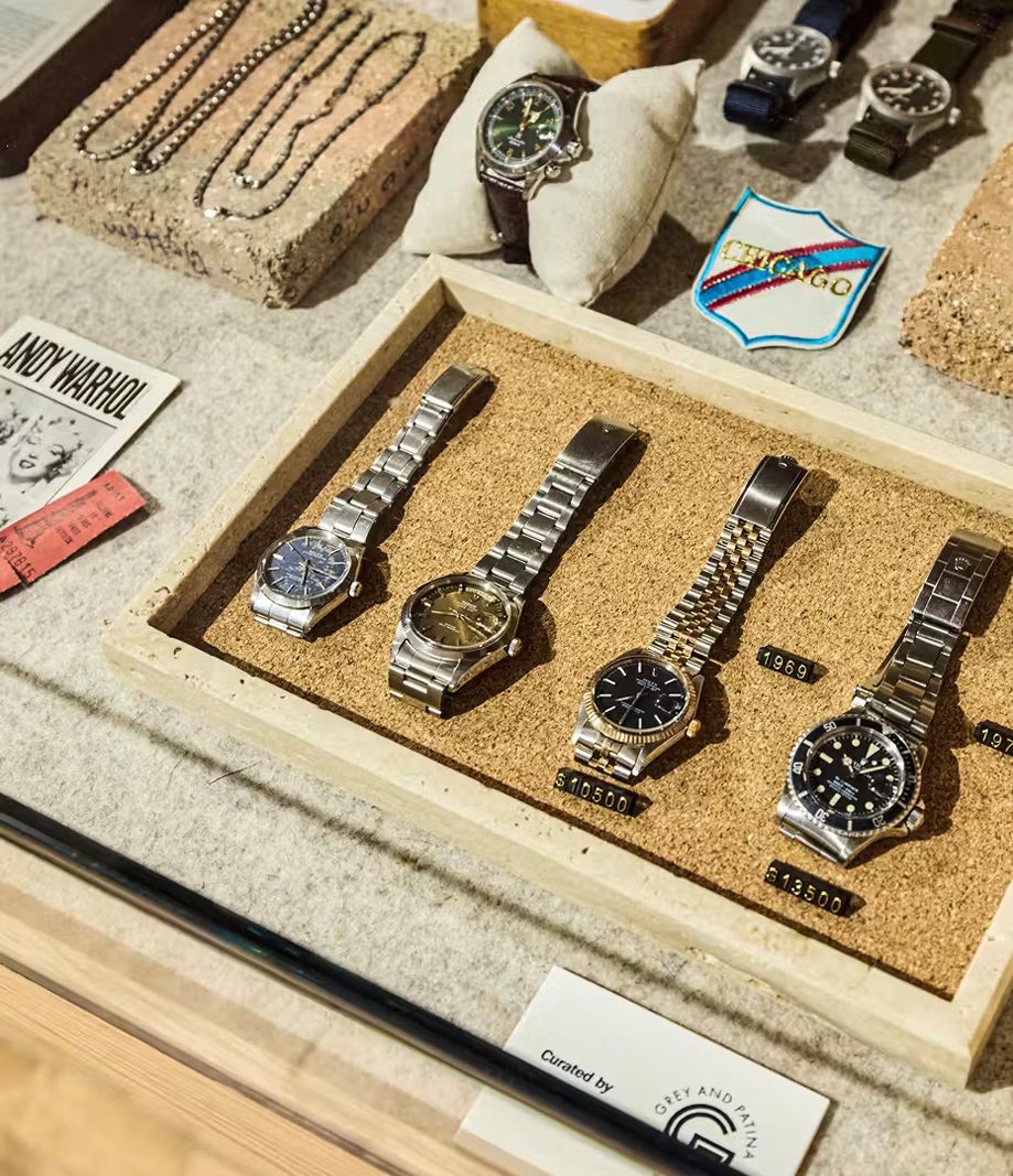 Display of wristwatches and necklaces arranged on cork-lined trays, with labels and a Curated by Grey and Patina card.