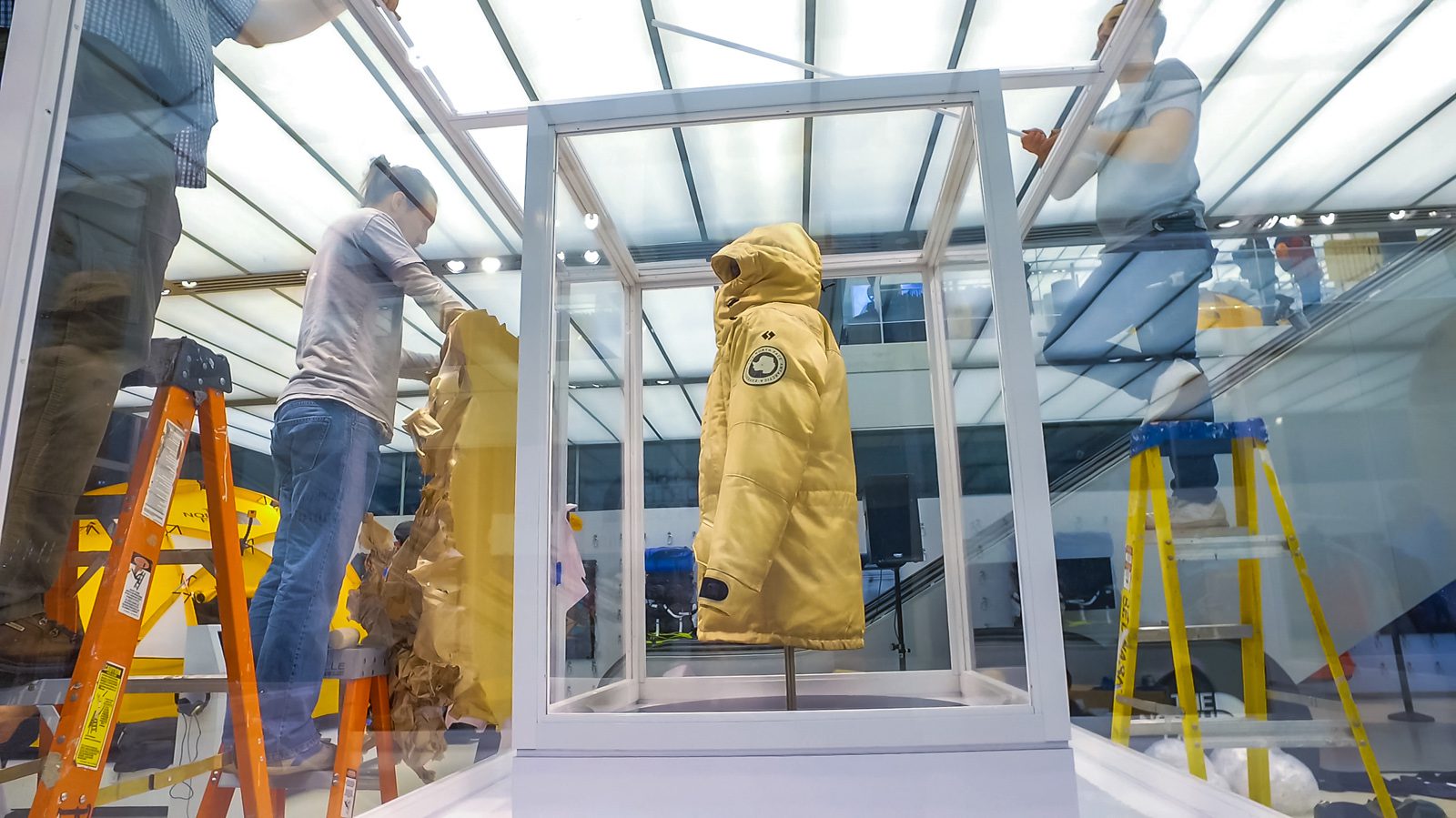 Yellow hooded jacket displayed in a glass case while workers set up the surrounding retail display