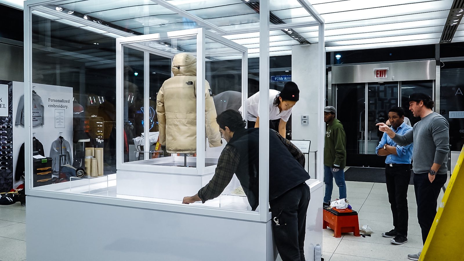 The North Face puffer jacket displayed in a glass case beside a personalized embroidery station and people working in the retail space.