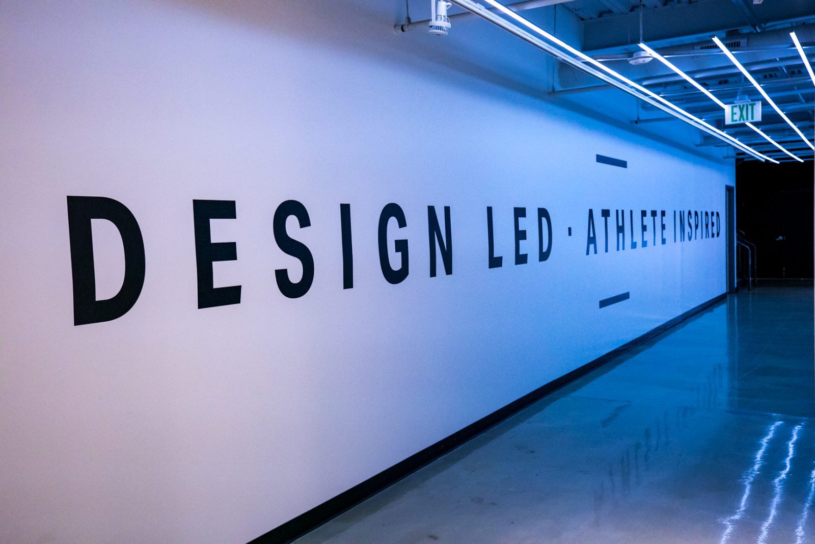 Blue-lit hallway wall with large text reading “DESIGN LED · ATHLETE INSPIRED” and an EXIT sign.