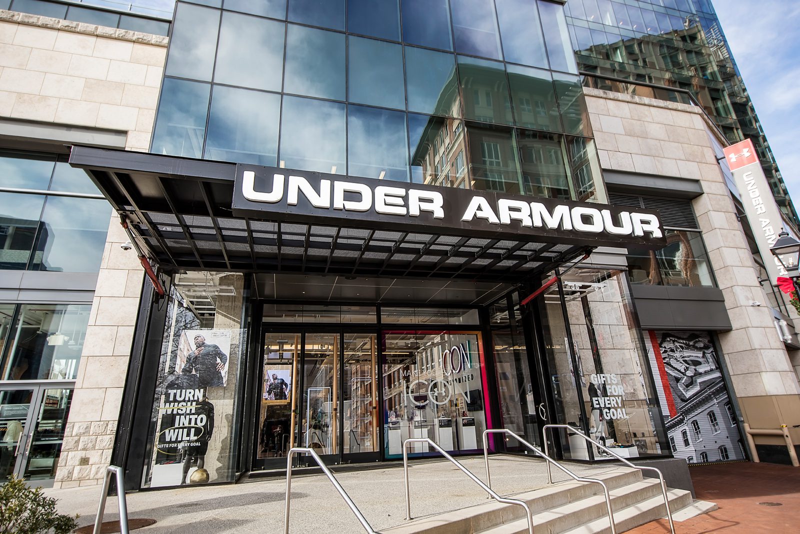 Exterior entrance of an Under Armour retail store with large Under Armour signage and promotional window displays.
