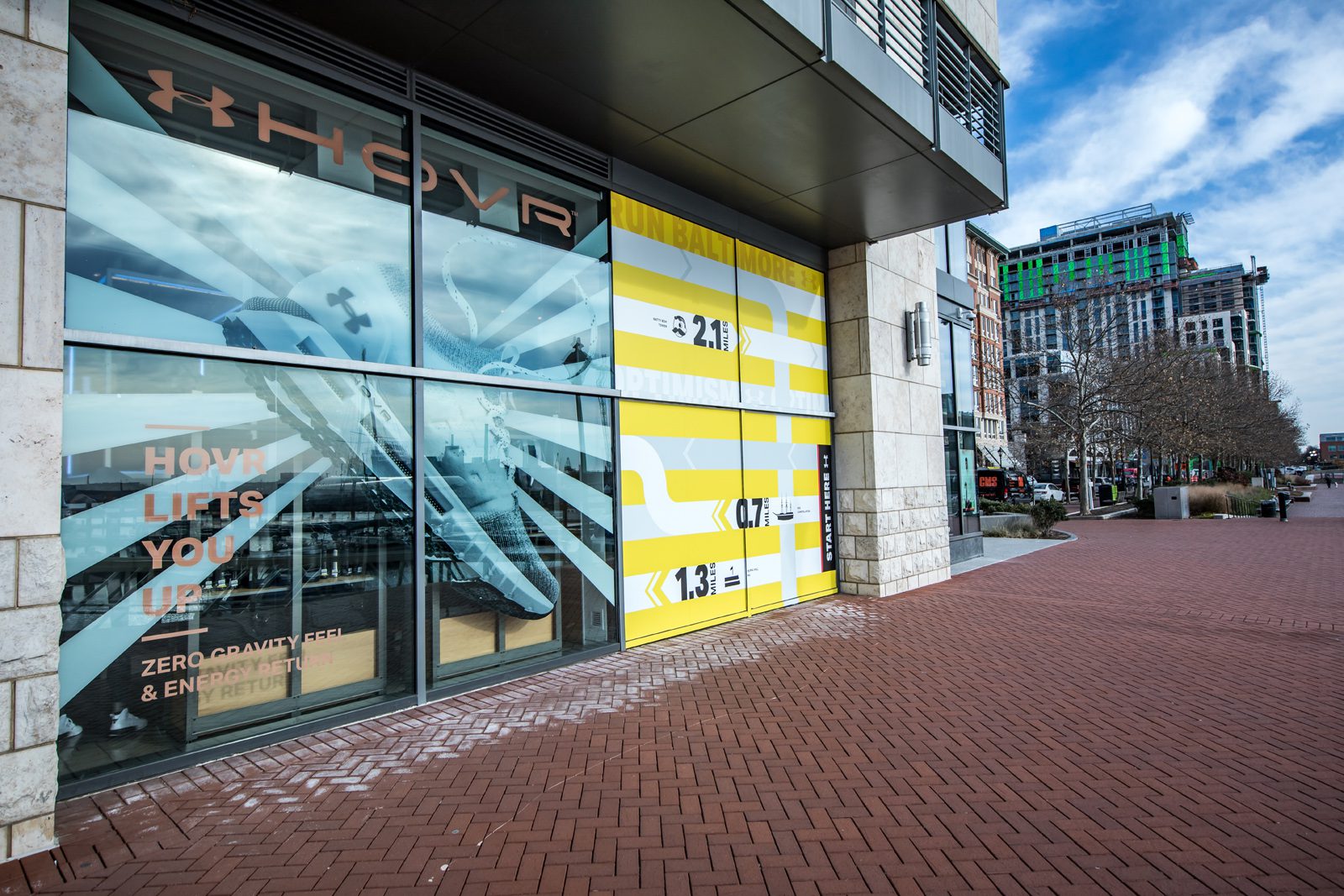 Under Armour storefront with yellow-and-white fitness campaign graphics and a brick-paved sidewalk in Baltimore