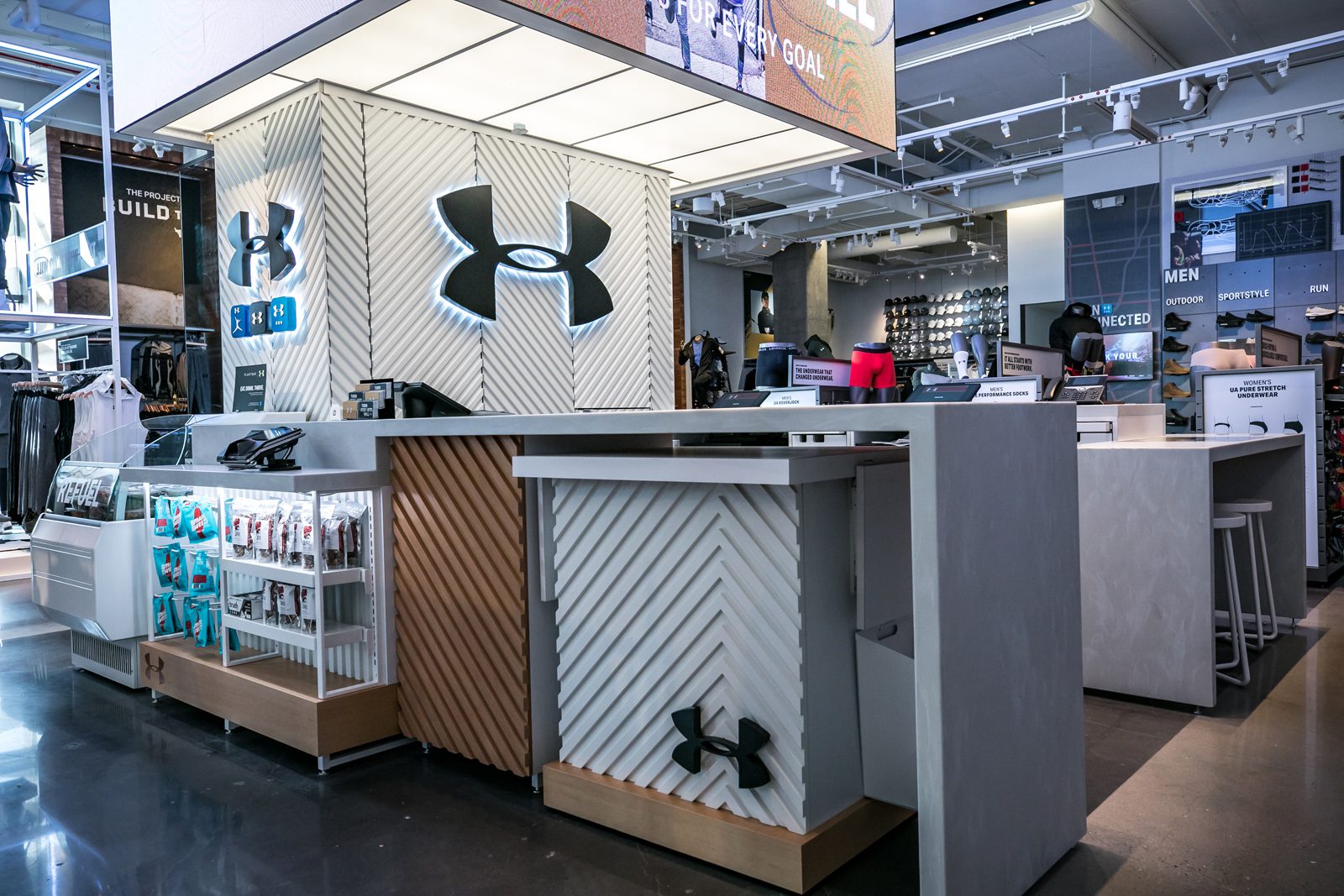 Interior of an Under Armour retail store with branded checkout counters, apparel displays, and sports equipment shelves
