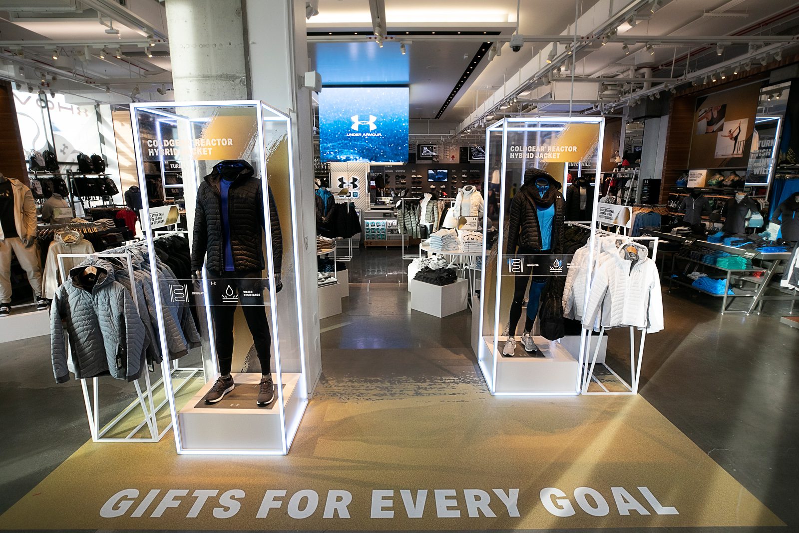 Under Armour store interior with clothing displays, mannequins, and floor text reading “GIFTS FOR EVERY GOAL.”