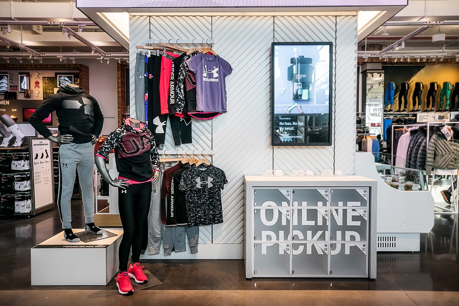 Under Armour store interior with athletic clothing displays, mannequins, footwear shelves, and a digital product advertisement.