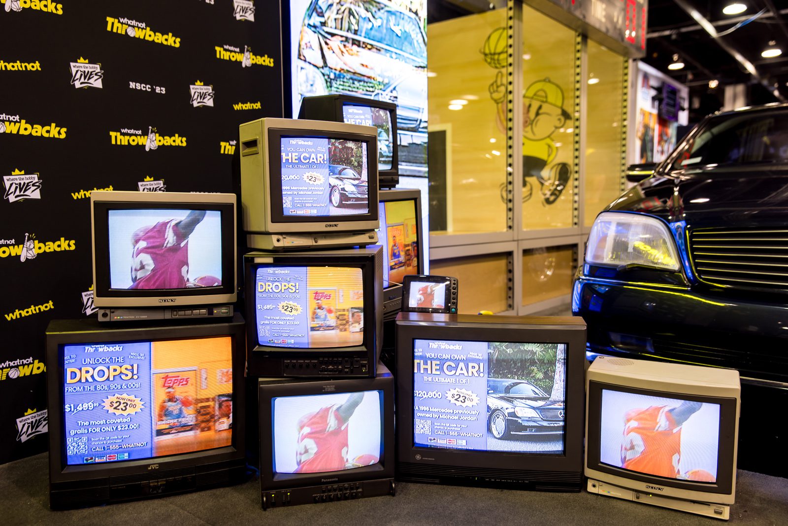Whatnot Throwbacks booth with stacked vintage CRT televisions showing promotional screens beside a black car, marked NSCC ’23.