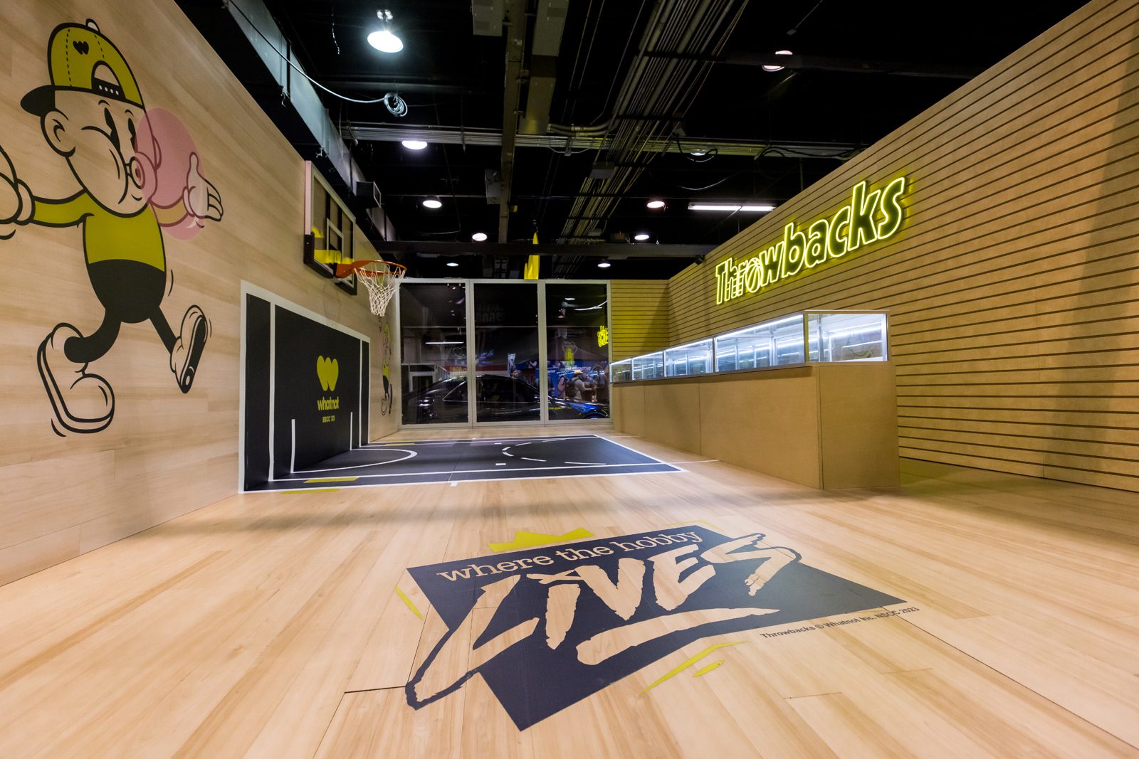 Throwbacks basketball-themed space with a wall mural, hoop, neon sign, display cases, and floor graphic reading “where the hobby lives.”