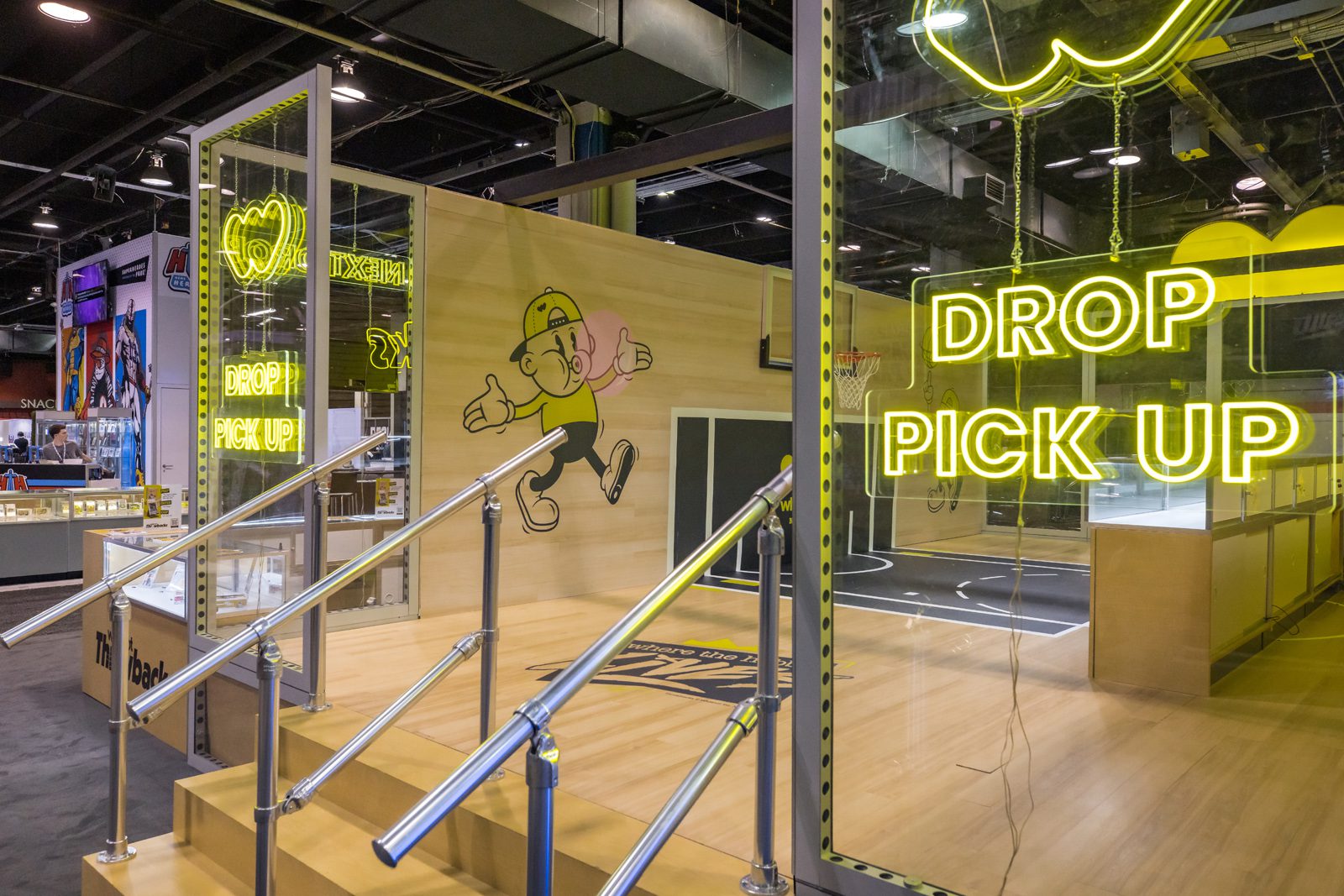 Whatnot event booth with neon “DROP PICK UP” signs, a cartoon mascot, display cases, and a small basketball court