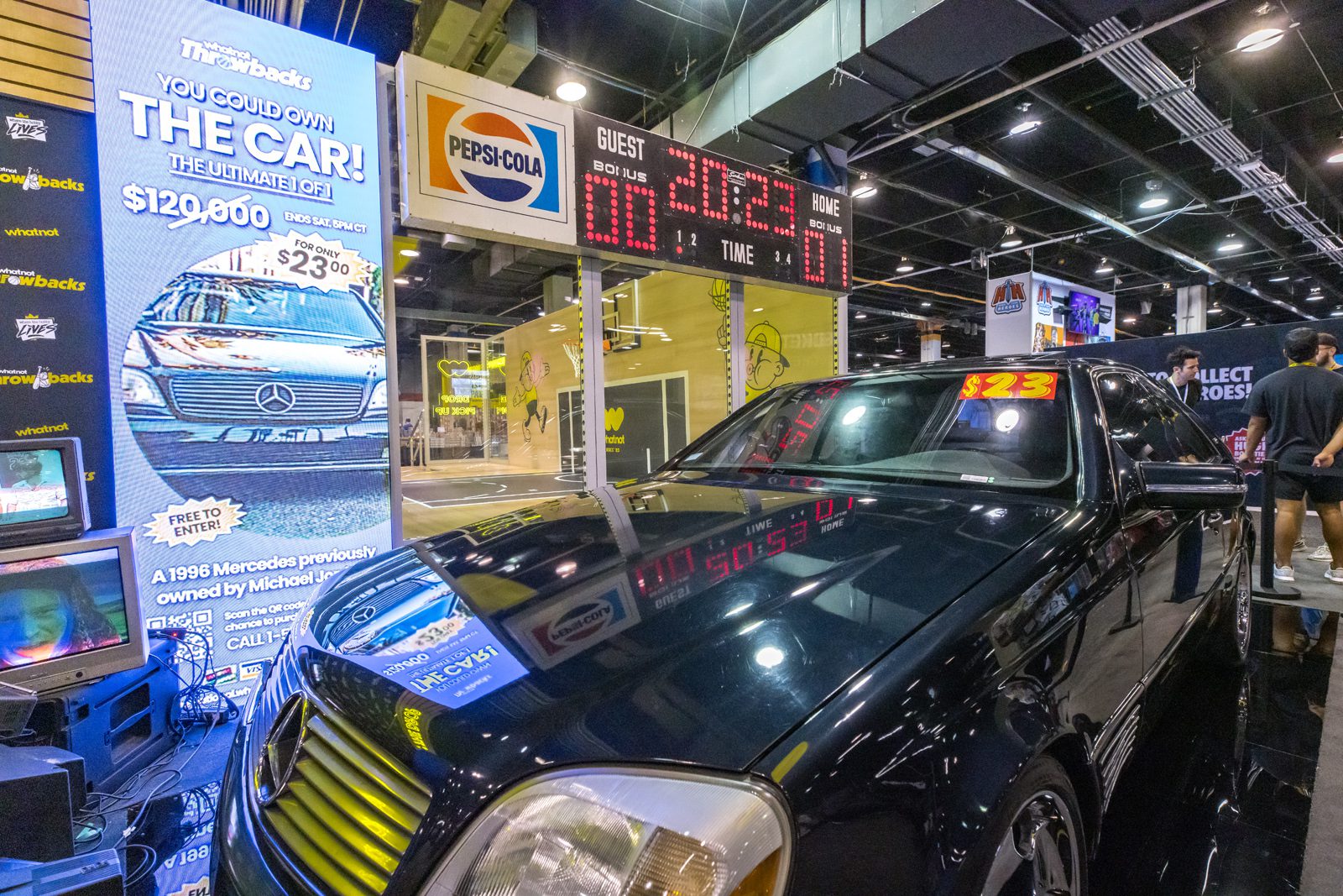 Black 1996 Mercedes displayed beneath a Pepsi-Cola scoreboard and promotional screen at a trade show.