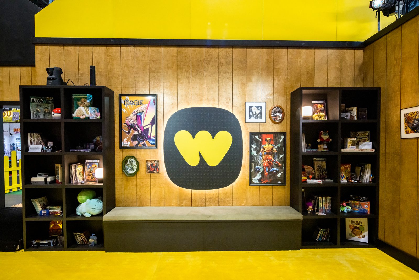Whatnot display wall with a large yellow logo, bench, and shelves of comic books, toys, and collectibles