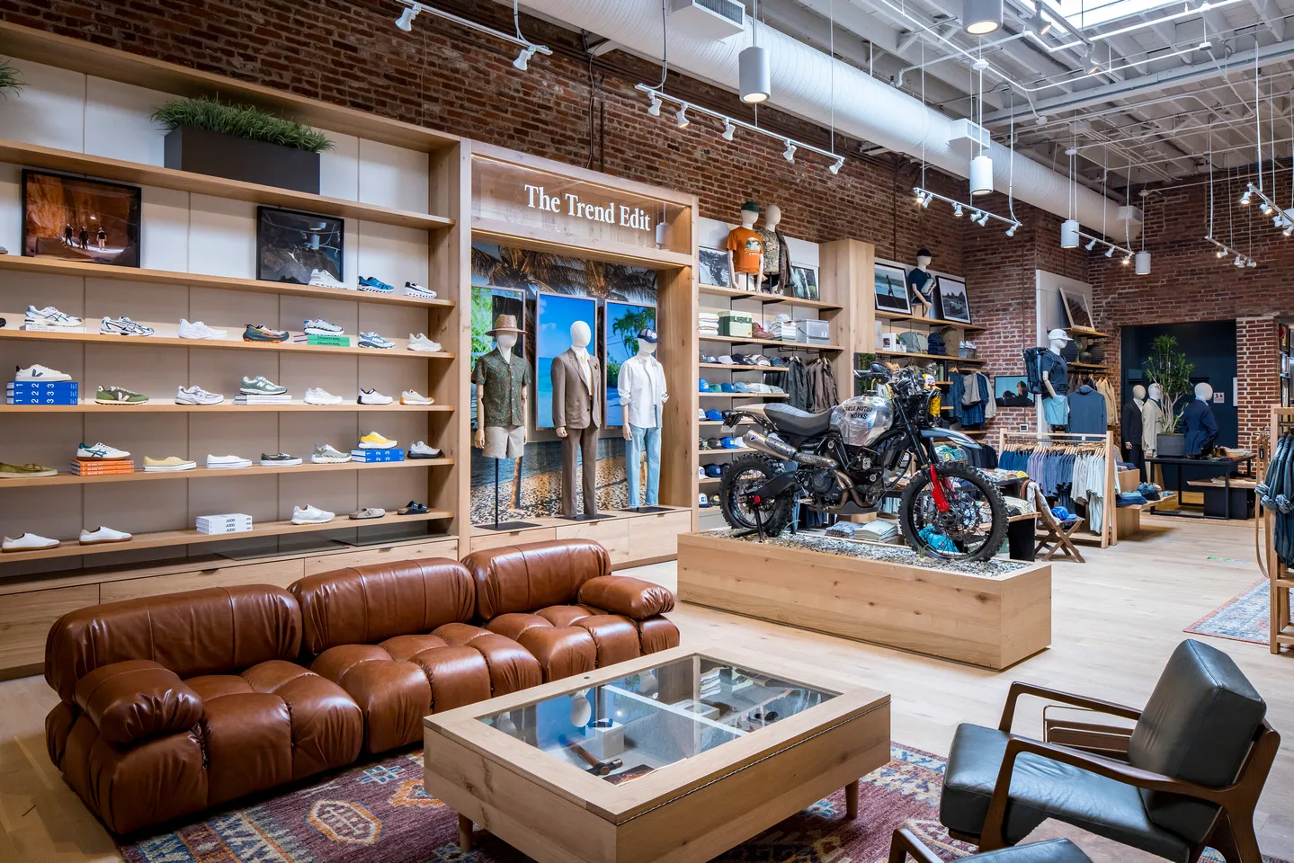 Retail store interior featuring clothing and shoe displays, a motorcycle on a platform, and a seating area beneath a “The Trend Edit” sign.