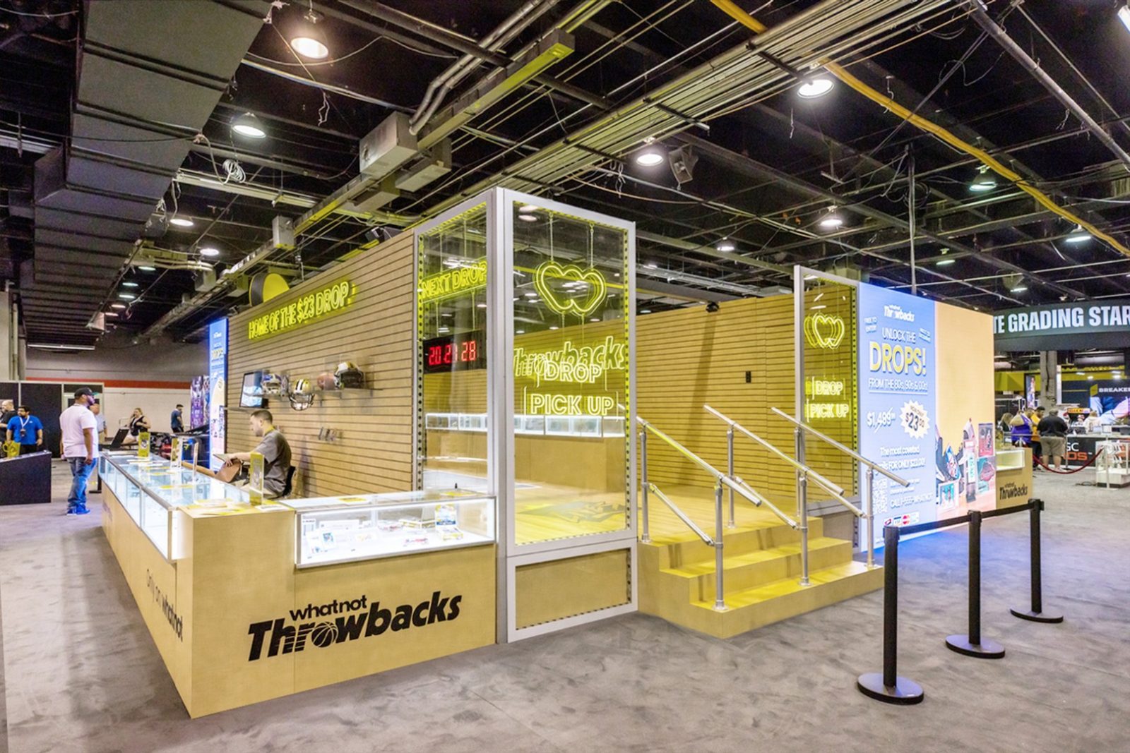 Whatnot Throwbacks booth with display cases, neon “DROP PICK UP” sign, stairs, and collectibles at a convention hall.