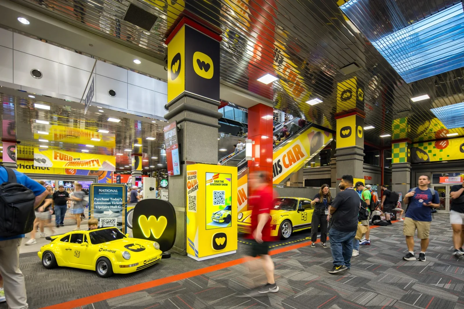 Whatnot-branded convention display with two yellow race cars, promotional signs, and attendees in a busy hall.