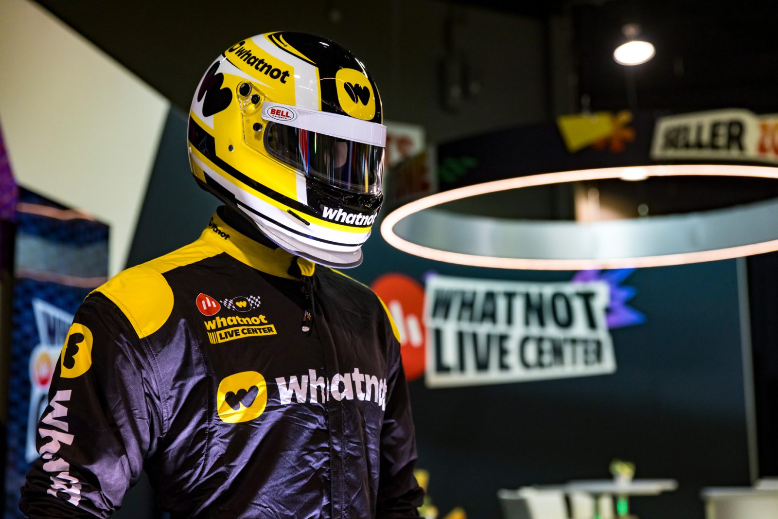 Whatnot-branded racing driver wearing a yellow-and-black helmet and suit beside a Whatnot Live Center display