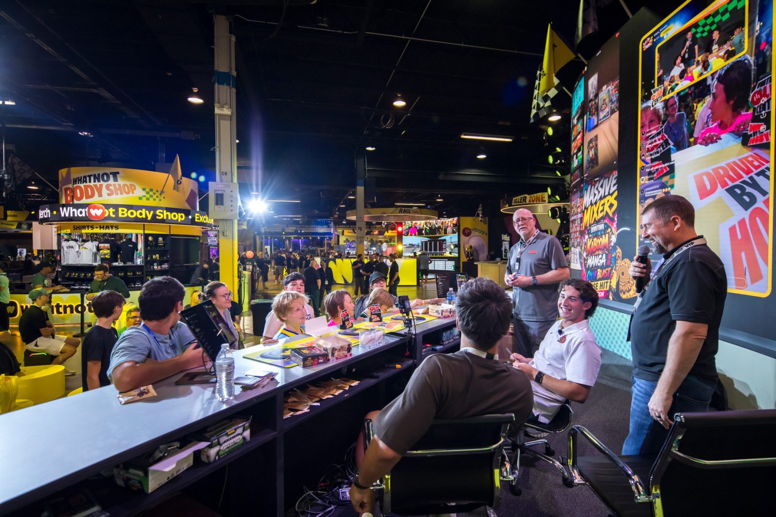 Attendees gather at a Whatnot Body Shop booth with merchandise, presenters, and large screens at a busy convention hall.