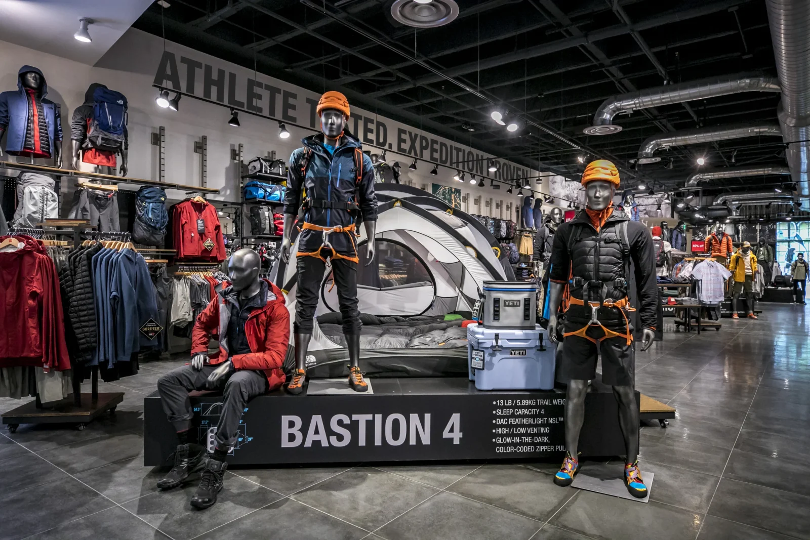 Outdoor gear store display featuring mannequins in climbing apparel, a tent, YETI coolers, and a Bastion 4 camping setup.