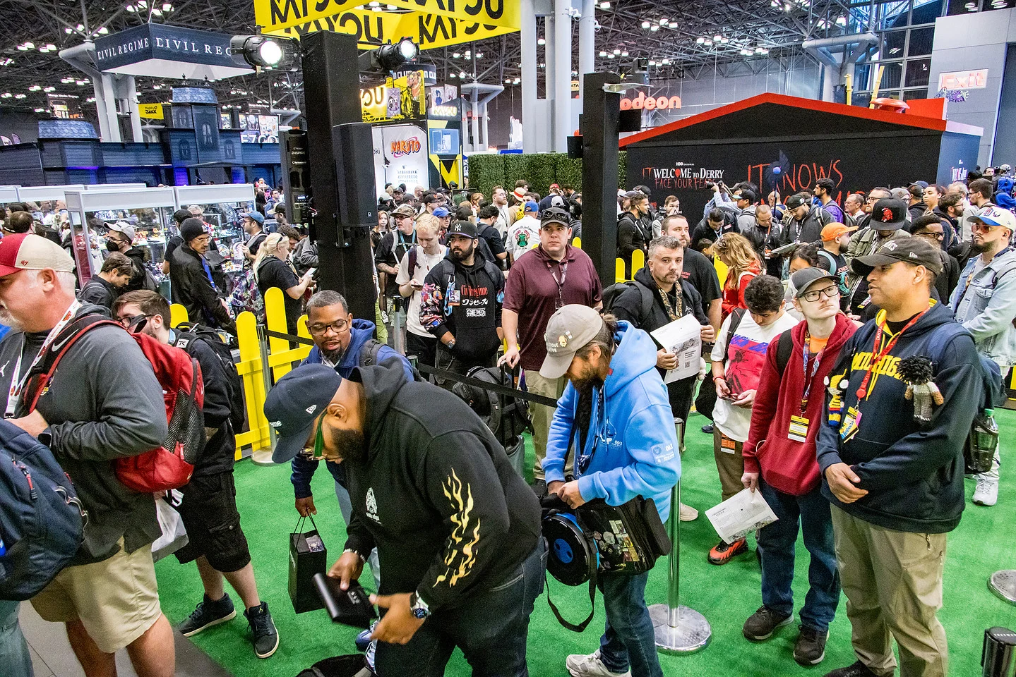 Crowded comic convention floor with attendees, vendor booths, queue barriers, and event signage
