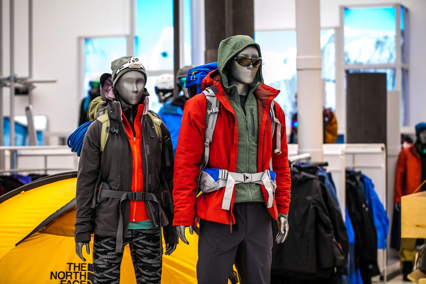 Two mannequins display layered hiking outfits with backpacks and sunglasses in an outdoor gear store.