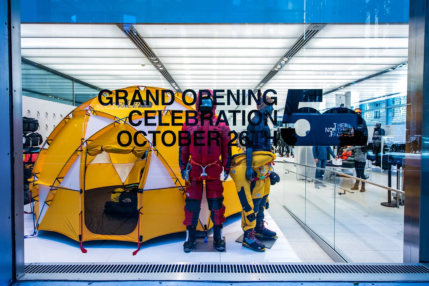 The North Face storefront display with yellow tents, climbing mannequins, and signage reading “Grand Opening Celebration October 26.”