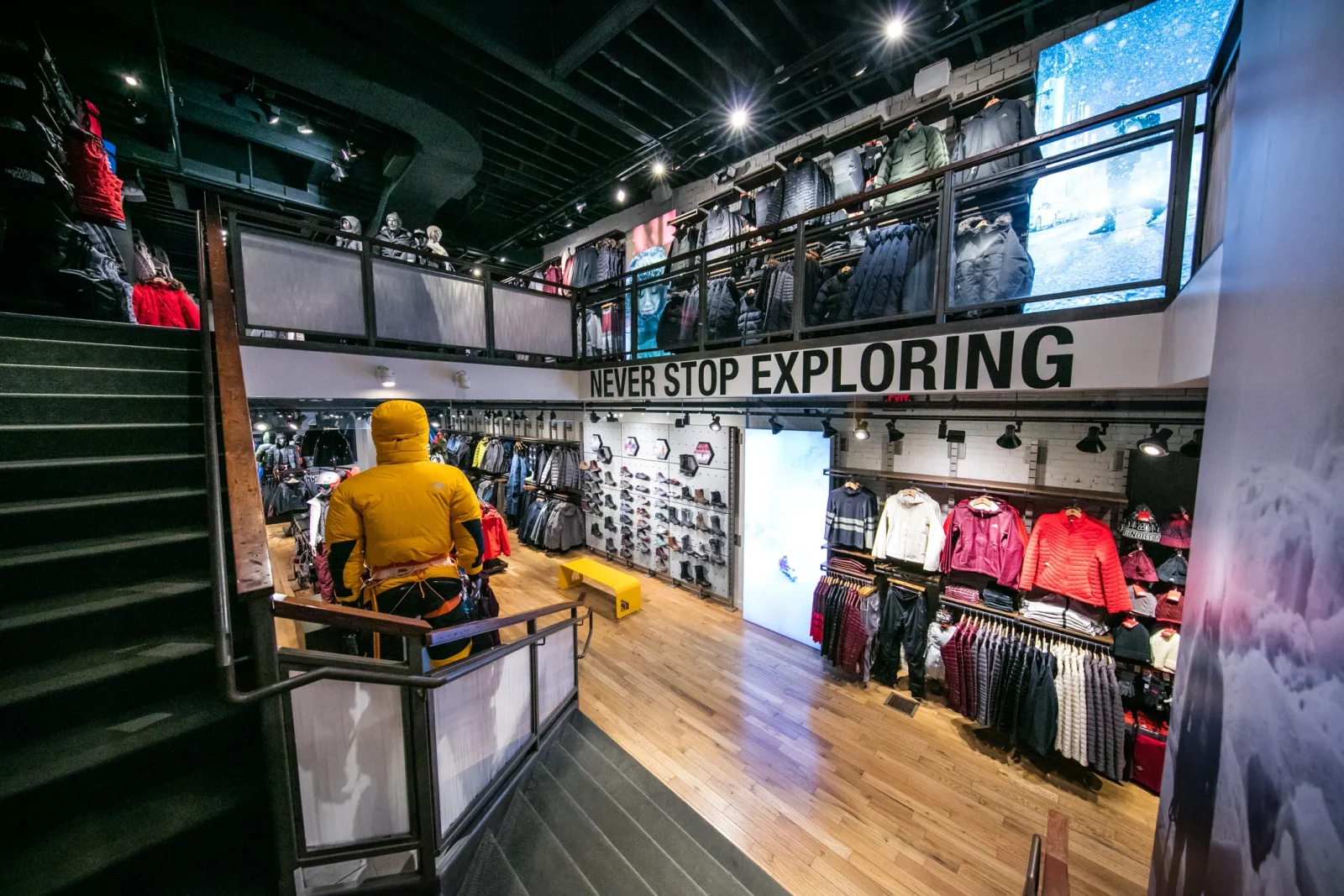 Interior of a two-level outdoor clothing store with jackets, footwear, mannequins, and a prominent “NEVER STOP EXPLORING” sign.