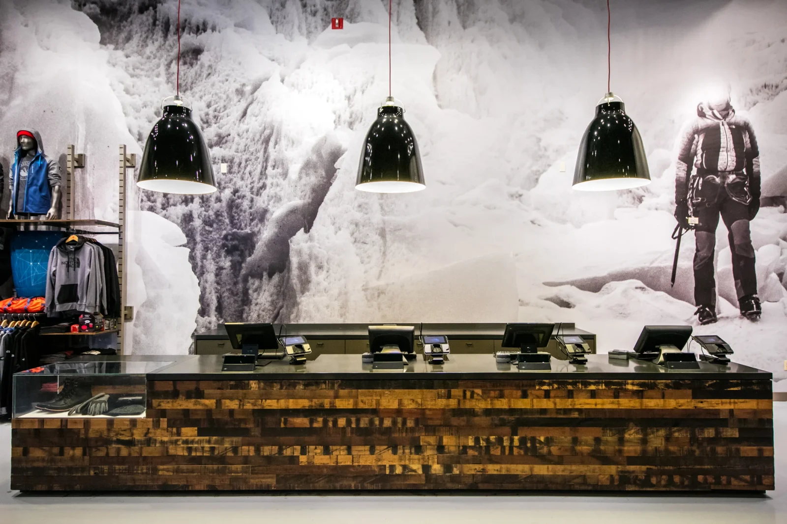 Outdoor apparel store checkout counter with clothing displays, pendant lights, and a snowy mountain-climbing mural