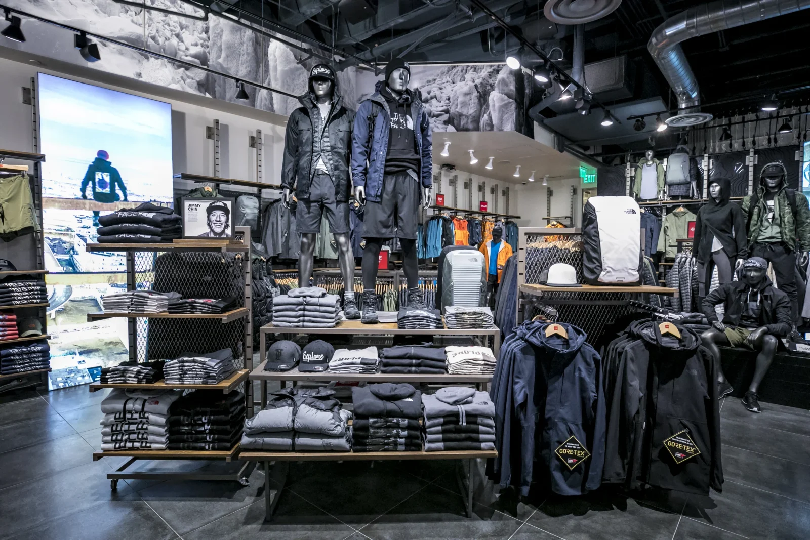 Outdoor apparel store interior with mannequins modeling jackets and shorts, folded clothing displays, backpacks, and Gore-Tex garments