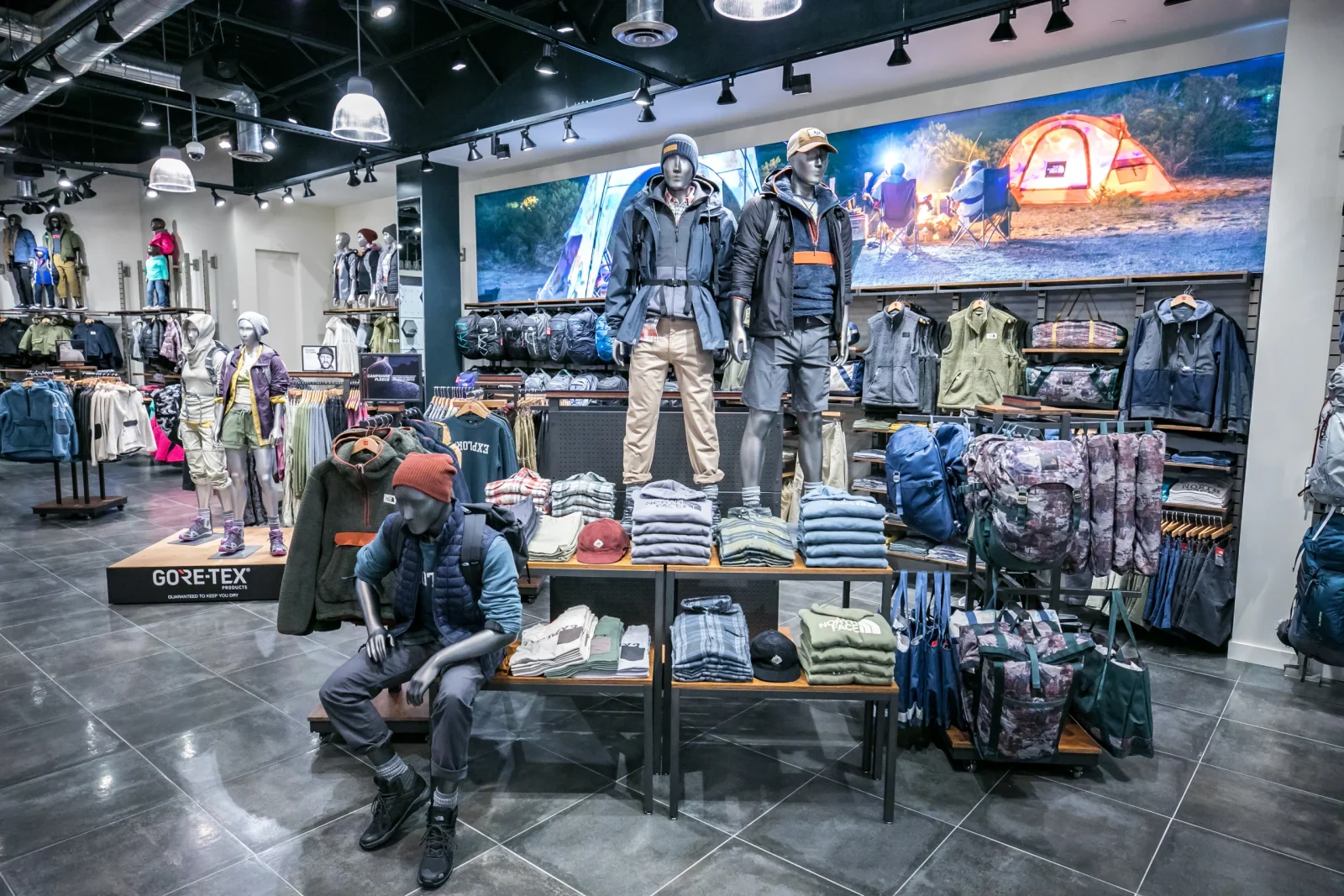 Outdoor apparel store interior with mannequins wearing jackets and hiking clothing, folded garments, backpacks, and camping gear on display.