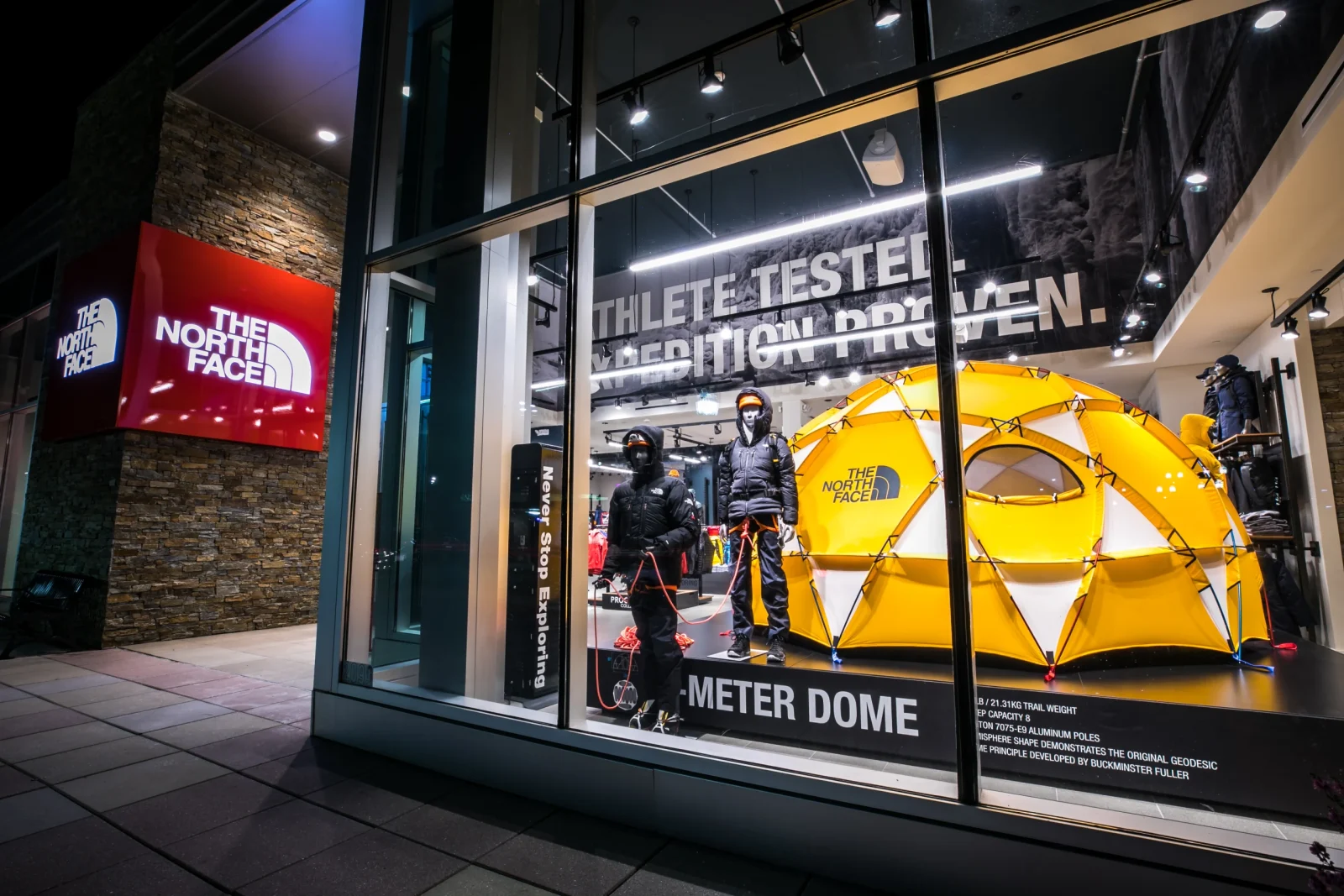 Outdoor apparel storefront window displaying mannequins in expedition clothing beside a bright yellow geodesic tent.