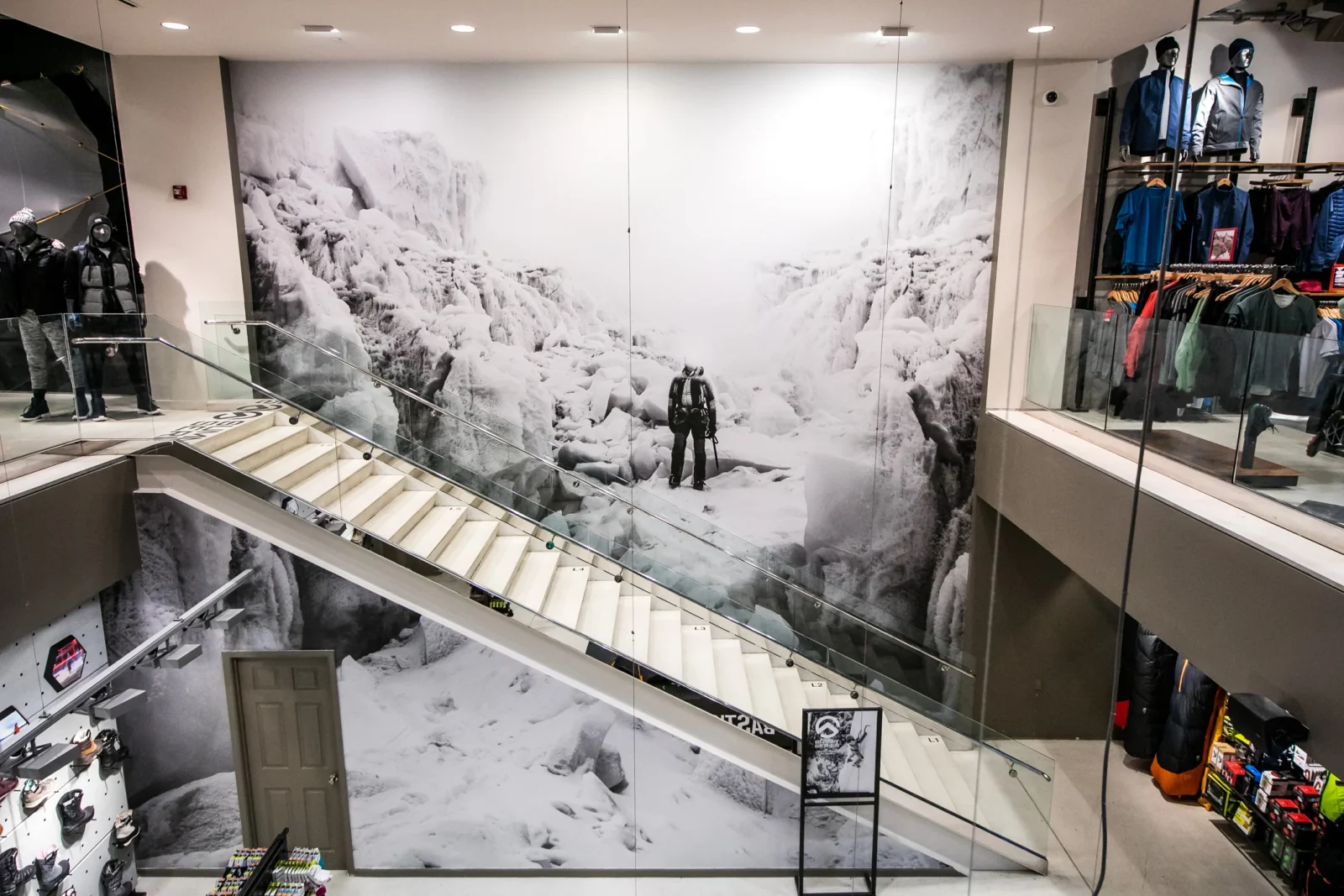 Outdoor clothing store interior with mannequins, apparel displays, a staircase, and a large snowy mountain mural
