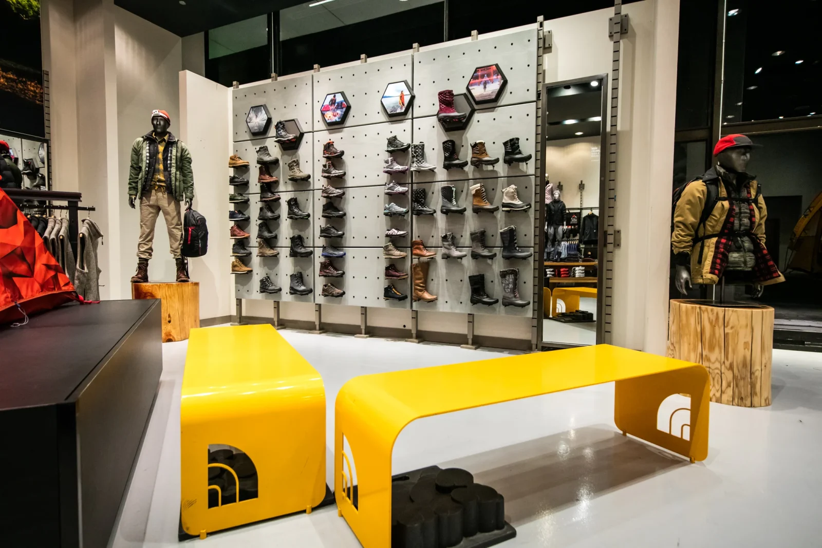 Outdoor retail store interior with mannequins, a wall of hiking footwear, camping gear, and bright yellow seating
