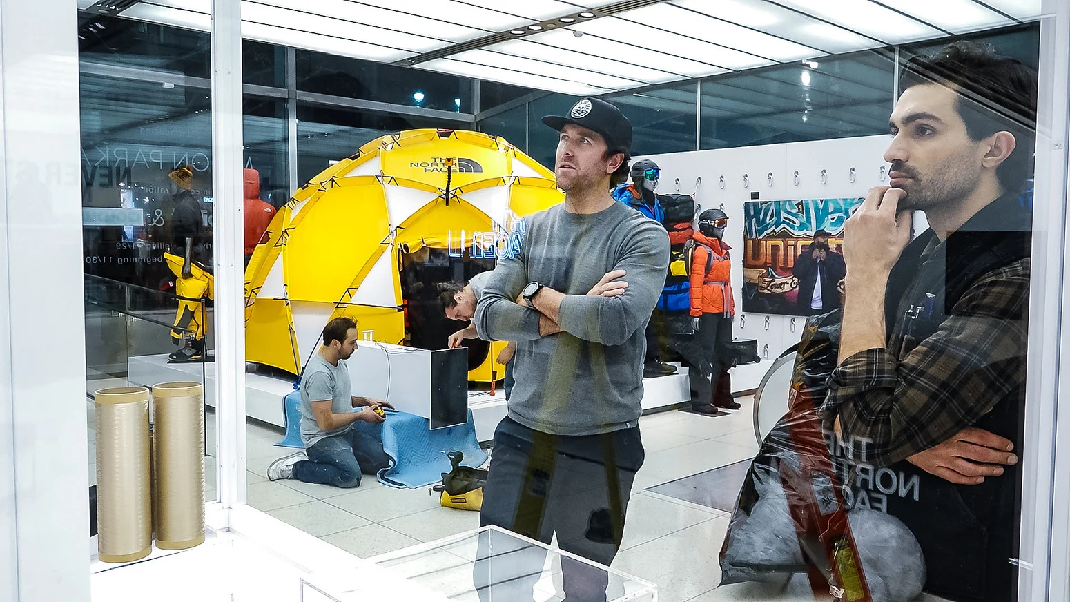 Outdoor gear shop display with a bright yellow tent, mannequins in jackets, and shoppers inside the store