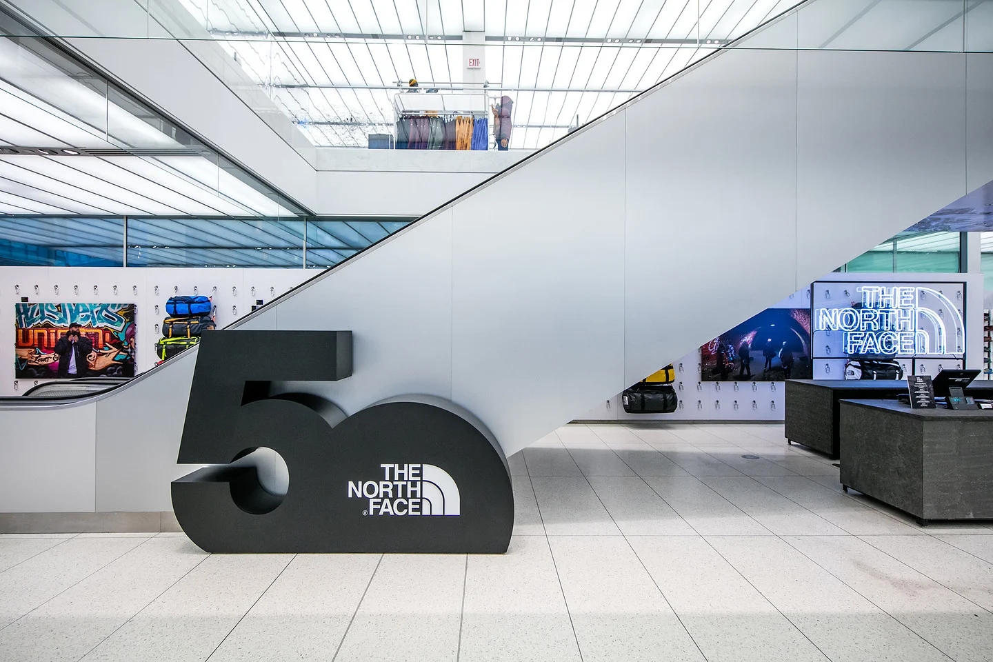 Interior of a The North Face store with a large branded display, escalator, outdoor gear, and illuminated The North Face sign.