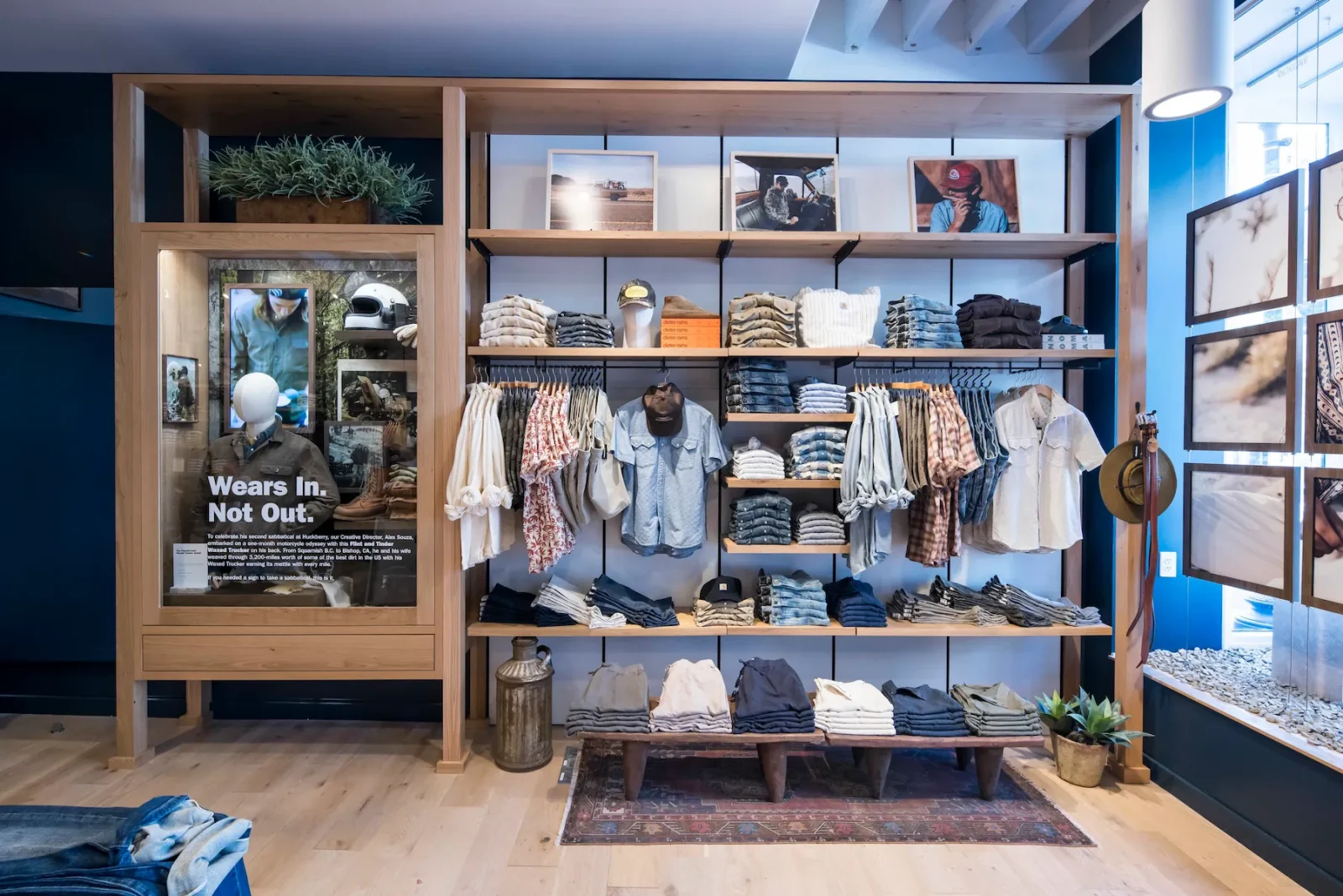 Retail clothing display with folded and hanging garments, framed photographs, and a mannequin behind a sign reading “Wears In. Not Out.”