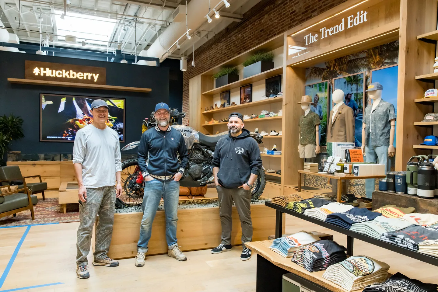 Huckberry store interior with three people, a motorcycle display, clothing tables, mannequins, and signs reading “Huckberry” and “The Trend Edit.”