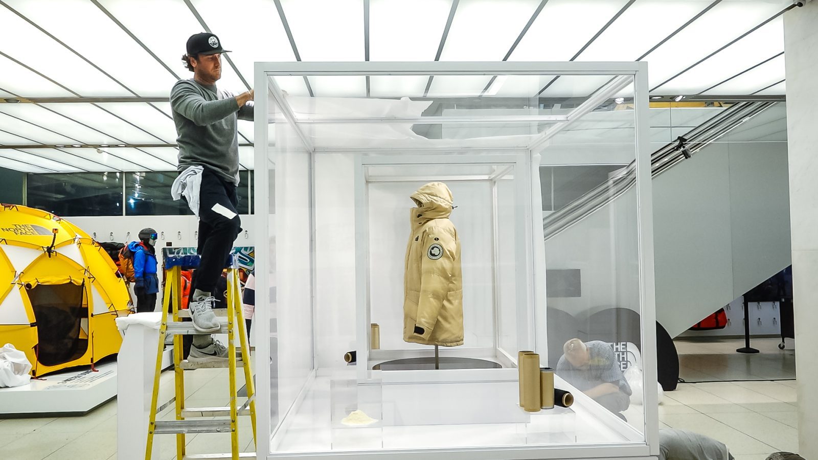 Worker installs a display featuring a beige hooded jacket inside a glass case, with a yellow tent and outdoor gear nearby.