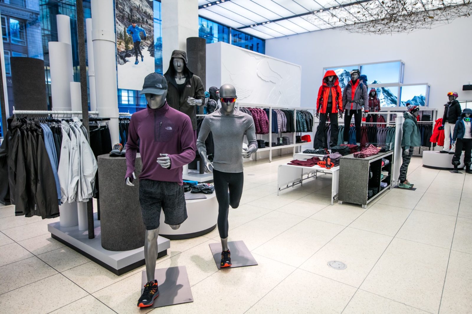 Outdoor apparel store interior with mannequins modeling jackets, athletic clothing, and winter gear alongside clothing racks and mountain displays.