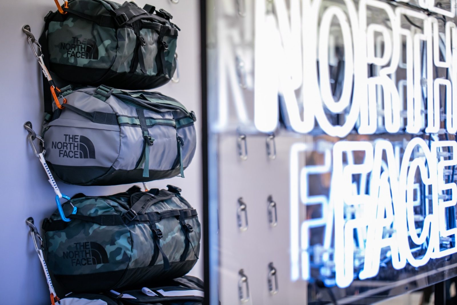 The North Face duffel bags displayed on a store wall beside a lit brand sign.