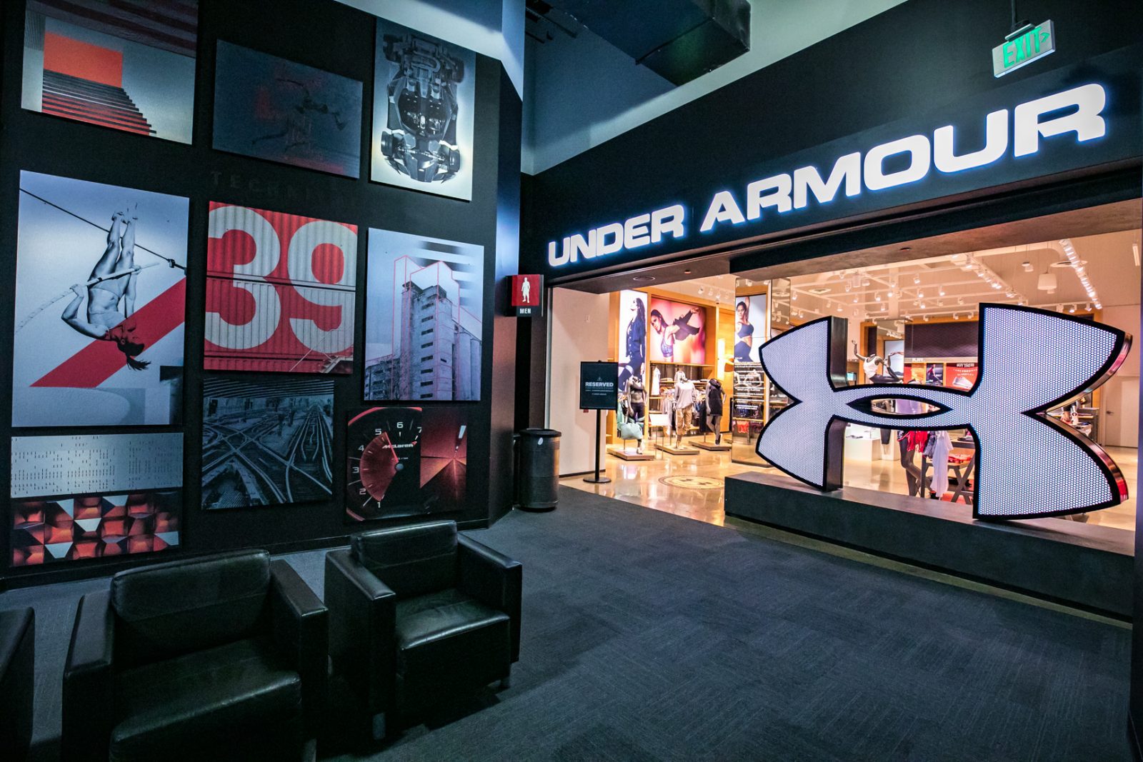 Under Armour store entrance with illuminated logo, clothing displays, wall artwork, and seating area