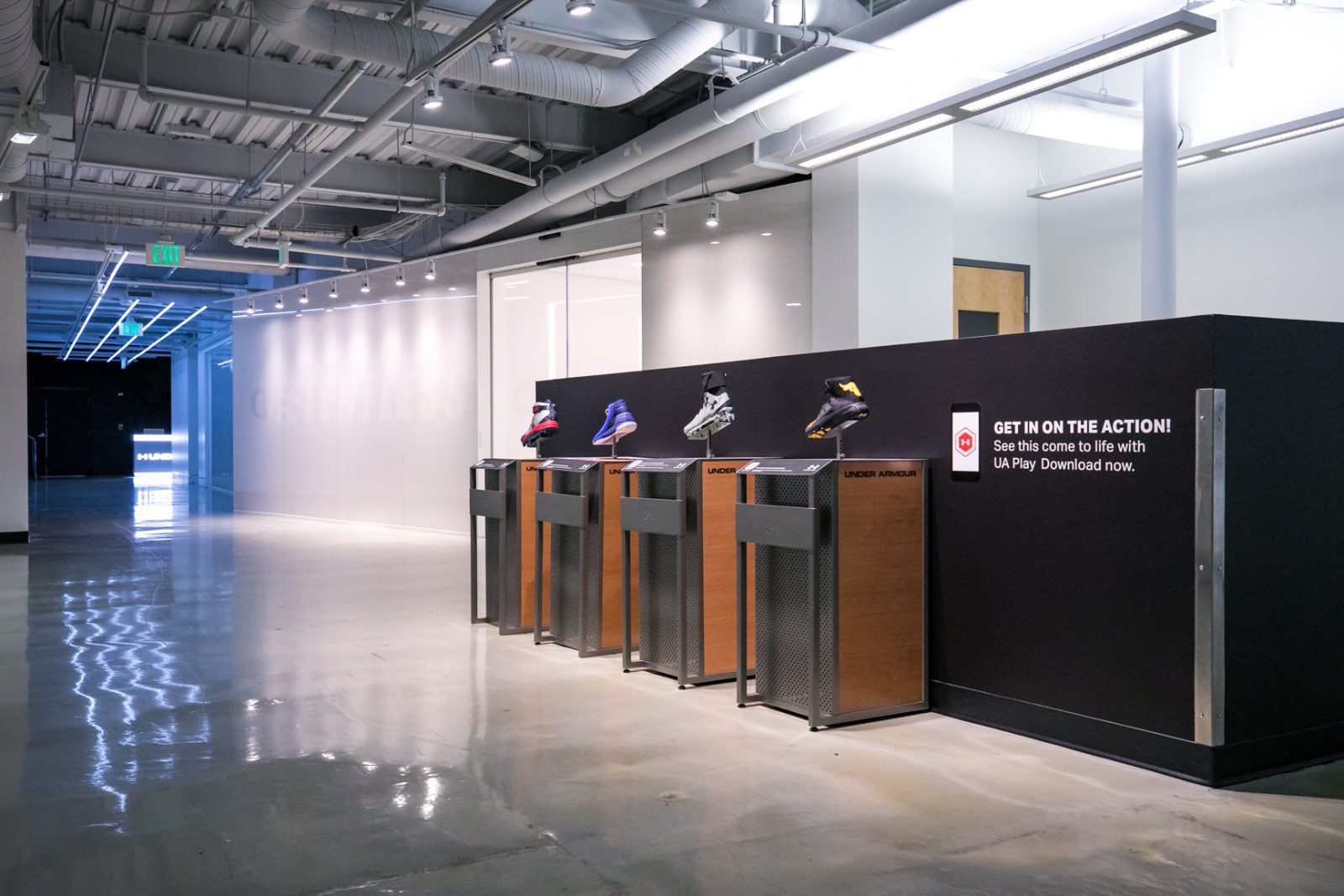 Under Armour showroom display featuring four athletic shoes and a sign promoting the UA Play app.