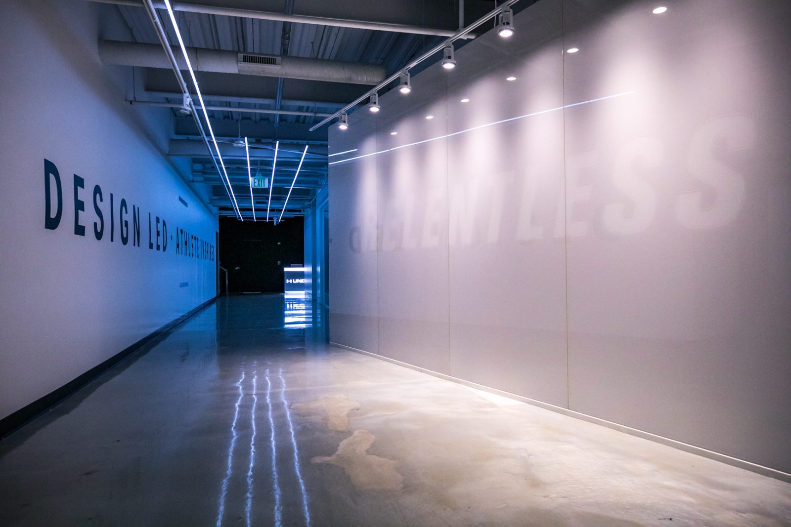 Blue-lit corridor with polished concrete floors, track lighting, and Under Armour branding on the walls.