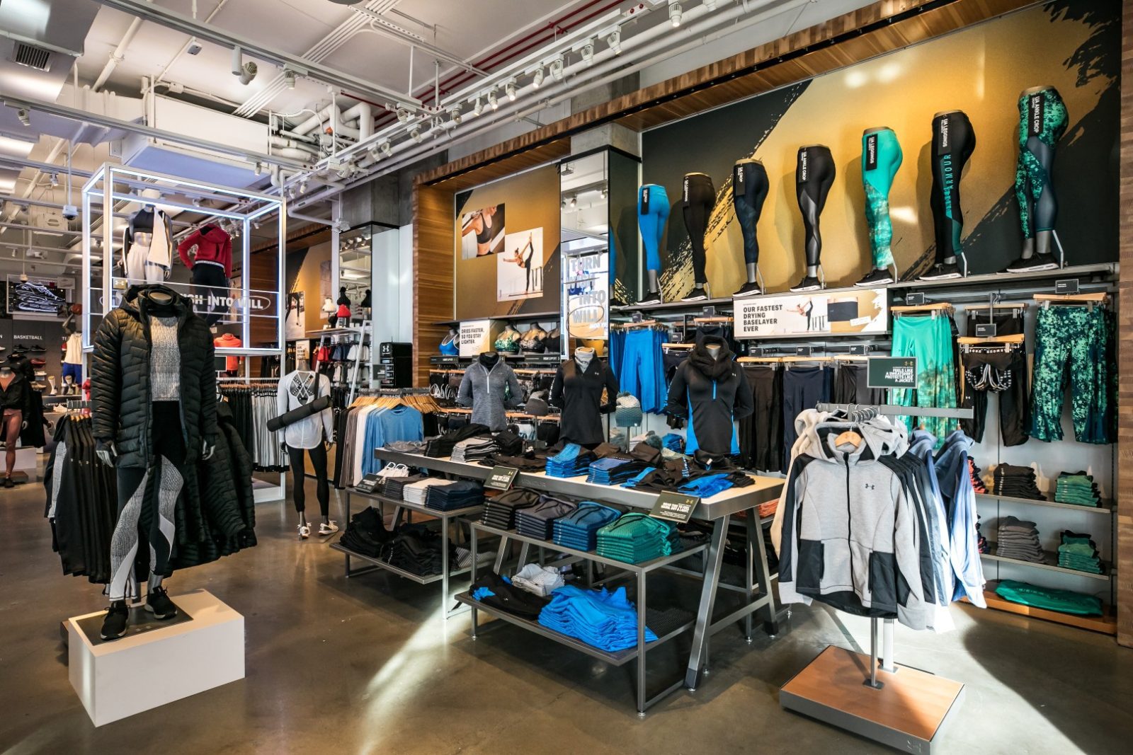 Interior of an Under Armour clothing store displaying athletic apparel, mannequins, clothing racks, and leggings on wall-mounted forms.