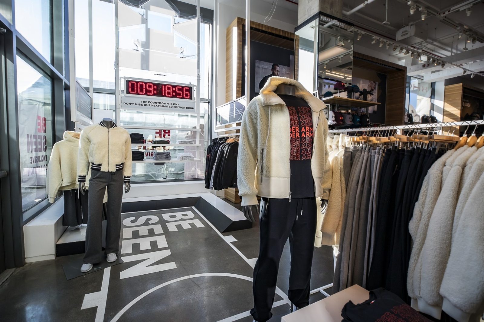 Under Armour store interior with mannequins in fleece jackets and athletic pants, clothing racks, and a countdown display for a limited-edition drop.