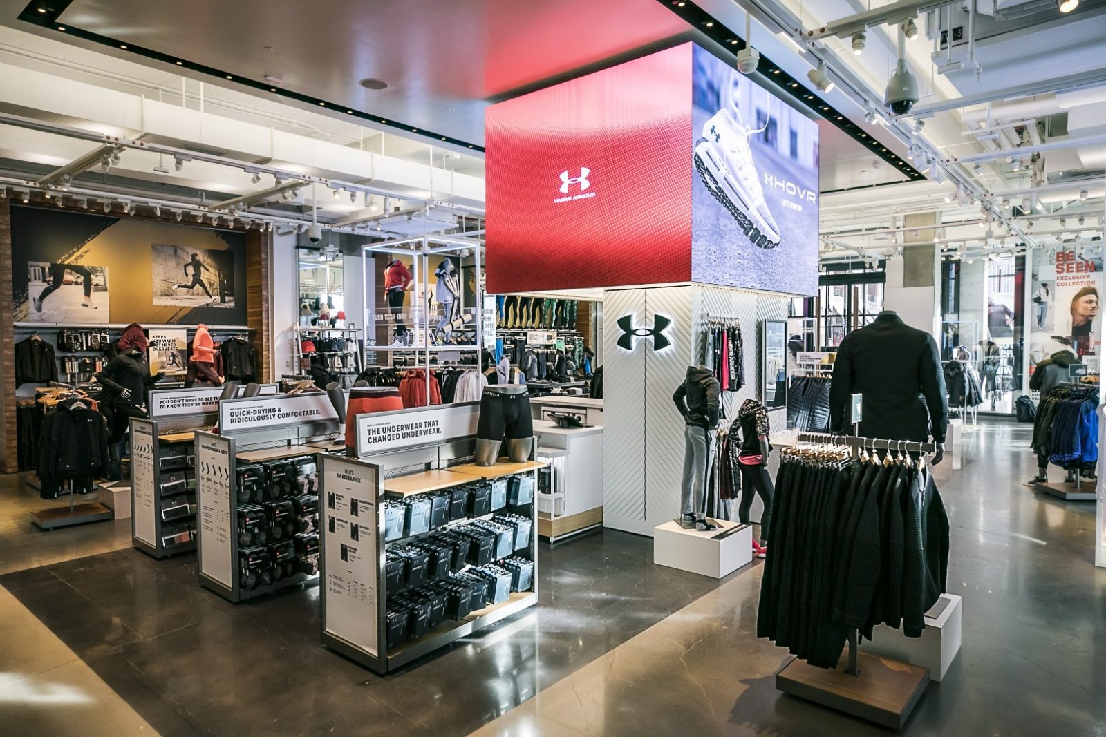 Interior of an Under Armour sportswear store with clothing displays, mannequins, product racks, and large digital advertising screens.