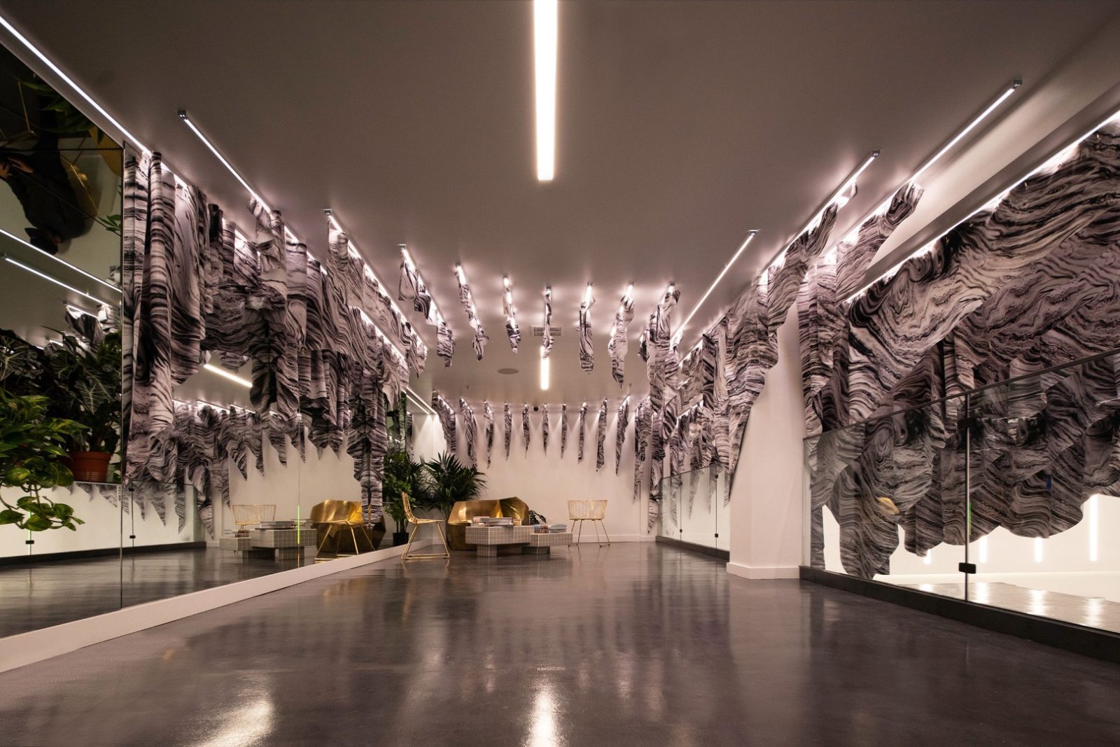 Interior space featuring draped marbled fabric, mirrored walls, modern seating, plants, and linear ceiling lights.