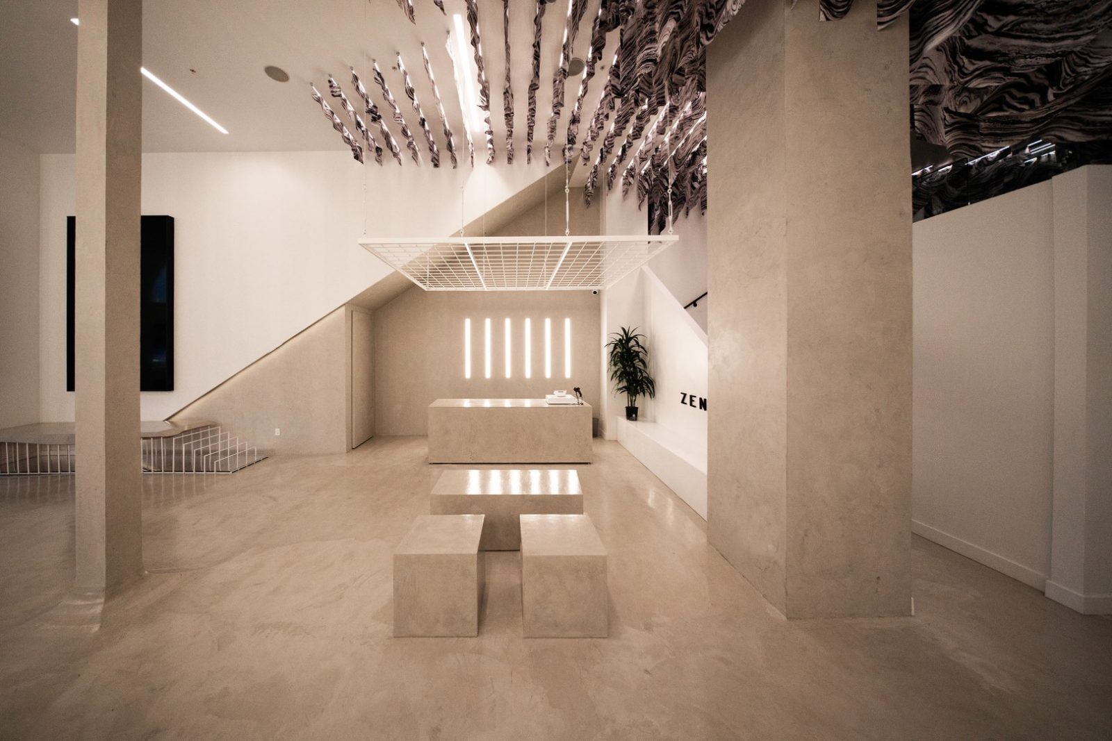 Minimalist Zensai store interior with a reception counter, geometric seating, staircase, plant, and sculptural ceiling installation