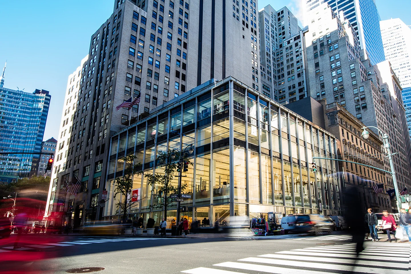Glass-fronted commercial building at a busy city intersection surrounded by tall skyscrapers, traffic, pedestrians, and an American flag.