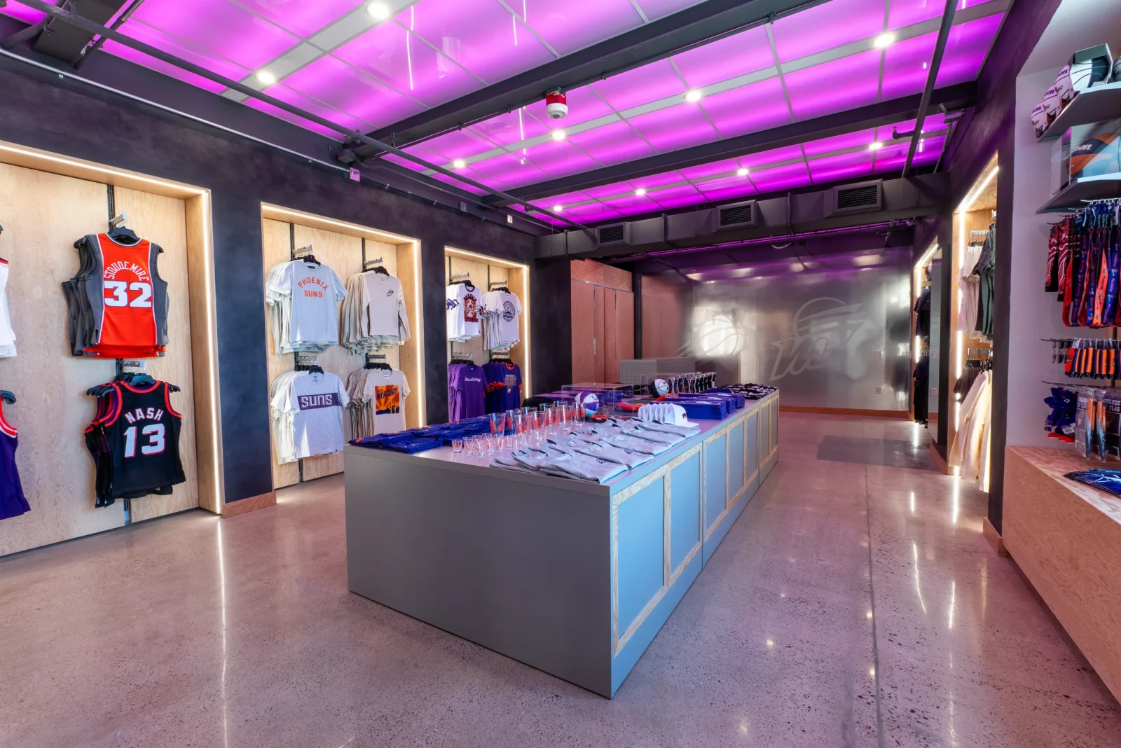 Interior of a Phoenix Suns team store displaying jerseys, shirts, hats, and basketball merchandise under purple lighting.