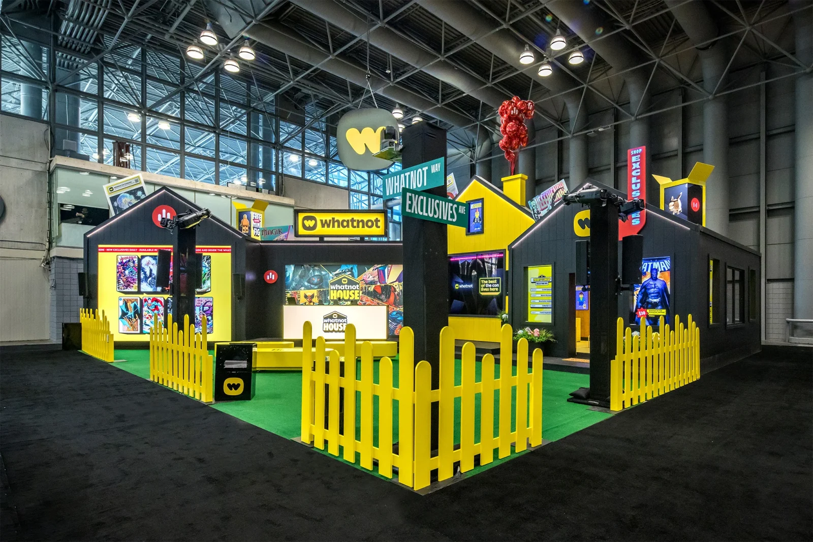 Whatnot House exhibition booth with black-and-yellow storefronts, comic displays, a yellow picket fence, and signs reading Whatnot and Exclusives.